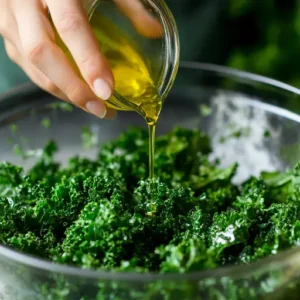 Hands massaging kale leaves with olive oil in a clear bowl for salad preparation.