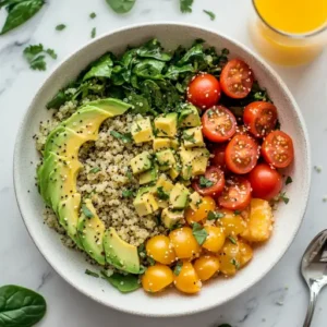 A colorful vegan quinoa breakfast bowl with avocado, cherry tomatoes, spinach, and nutritional yeast on a white plate.